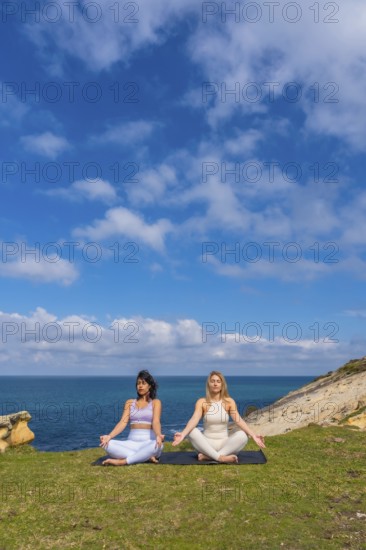 Two women practicing meditation and yoga in a serene lotus pose on a yoga mat overlooking the ocean from a grassy cliff, finding peace and wellness in nature