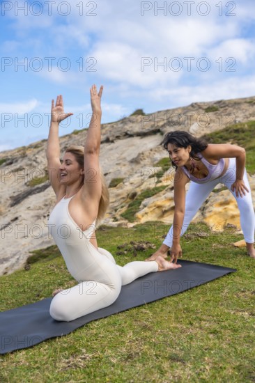 Two women practicing yoga together outdoors on a green grassy hill, one woman demonstrating a pigeon pose with arms raised while a female instructor helps adjust her foot