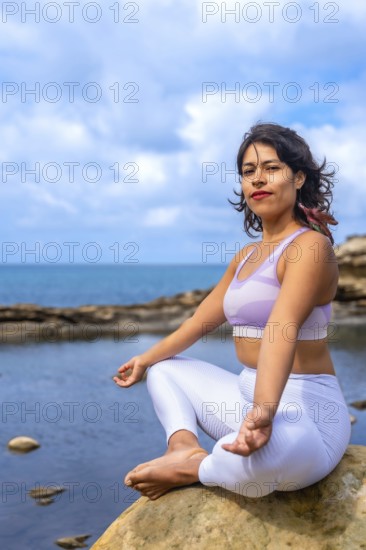 Woman sitting in a lotus position on a rock by the ocean, practicing yoga and finding inner peace, connecting with nature for wellness and spiritual balance