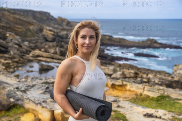 Blonde woman holding a yoga mat on a rocky coast, standing and preparing for outdoor meditation and stretching with the calm ocean and expansive coastline as backdrop