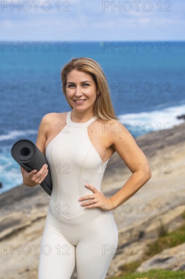 Woman smiling, looking at camera, holding yoga mat, ready for a yoga or meditation session outdoors, connecting with nature and enjoying a healthy lifestyle by the sea