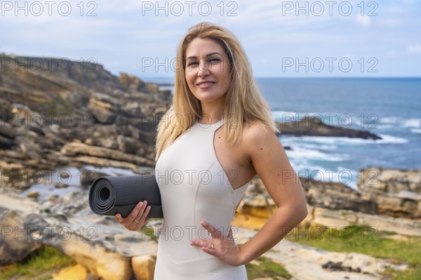 Woman stands on rocky coast by the ocean holding a yoga mat, smiling at camera before a yoga and meditation session amid sunny seascape, wellness and mindfulness practice