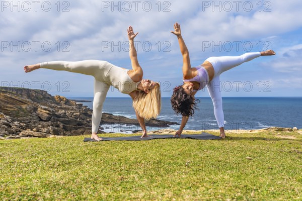 Two women performing a balancing yoga pose on mats on a grassy cliff overlooking the ocean, celebrating wellness, harmony, and connection with nature during an outdoor exercise session
