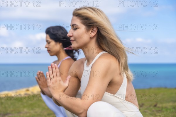 Two women with eyes closed practicing yoga and meditation in prayer pose on the seaside, embracing mindfulness, inner peace and spiritual wellness with ocean horizon and blue sky