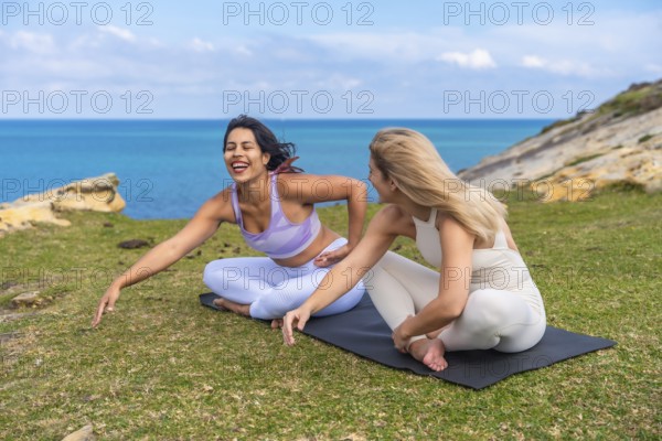 Two happy friends enjoying a yoga session and laughing together on a grassy cliffside overlooking the blue ocean, sharing a moment of wellness and friendship