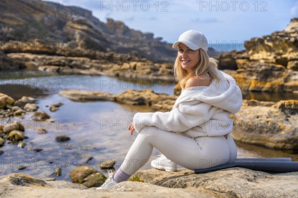 Blonde woman on a yoga mat by a calm tidal pool, sitting and smiling over her shoulder, ready for a mindful yoga session in a peaceful coastal natural setting