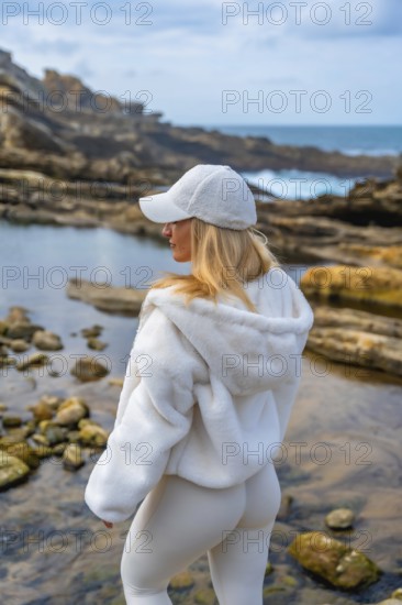 Woman in comfortable hoodie and leggings meditating on rocky coast, back to camera, finding calm and mindfulness beside the ocean under a tranquil sky, peaceful solitude