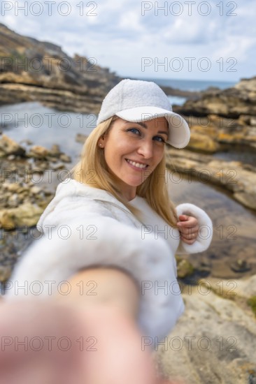 Blonde woman wearing a white cap and hoodie reaching out to take a selfie, smiling to the camera while standing on a rocky coast with tidal pools and the sea in the background