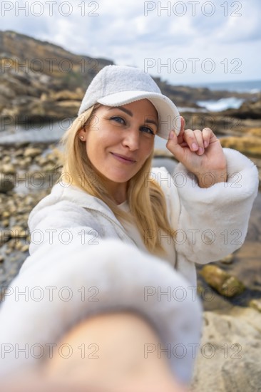 Happy woman smiling to camera and adjusting her white cap, enjoying a casual outdoor moment on a rugged coastline with a cloudy sky and rocky formations