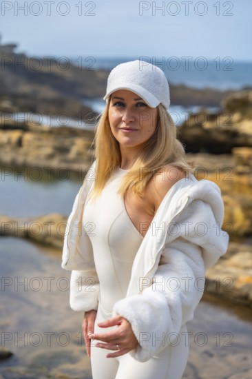 Woman in white sportswear and baseball cap stands on rocky shoreline by the blue sea, smiling confidently in sunny, relaxed outdoor fashion portrait evoking wellness and summer freedom