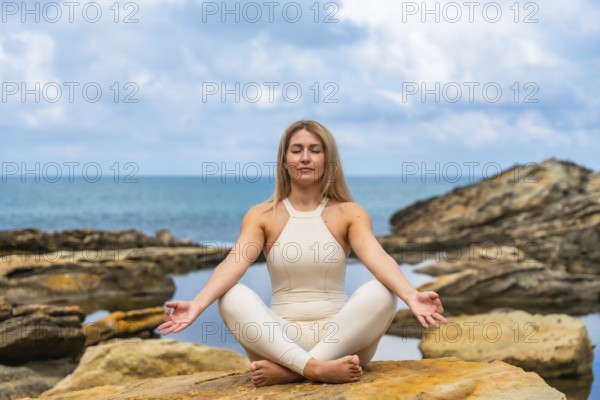 Woman practicing yoga outdoors, sitting on rocks in a peaceful lotus pose with eyes closed, finding calm and concentration by the ocean for wellness and spiritual balance
