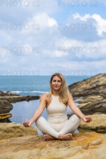 Woman in a white outfit sitting in a lotus position on a rock by the ocean, meditating and finding inner peace, embodying wellness, balance, and mindfulness in nature