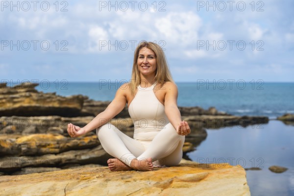 Woman practicing yoga in lotus on a rocky ocean cliff, meditating with eyes closed, embracing balance, calm and strength amid sea breeze and blue sky serenity