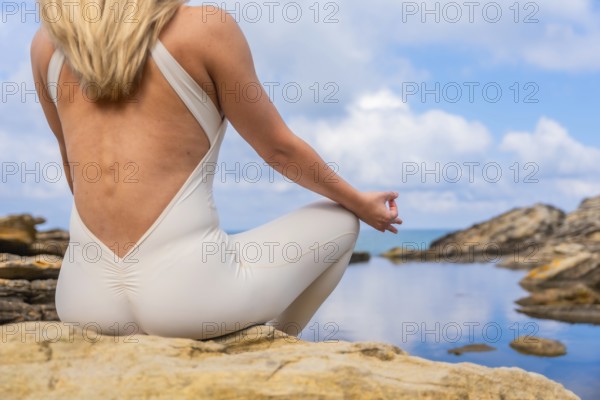 Woman practicing yoga and meditation in a peaceful outdoor setting, sitting in lotus pose on rocks beside a calm natural pool and the sea, promoting relaxation and wellness