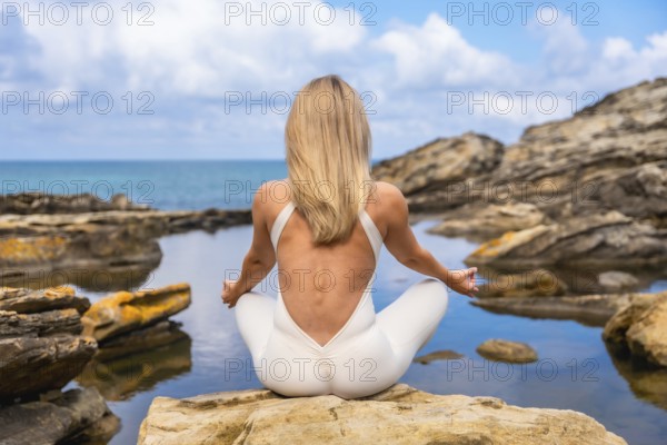 Woman in a white jumpsuit practicing a meditation yoga pose on coastal rocks, finding wellness and inner peace by the tranquil sea with a clear sky overhead