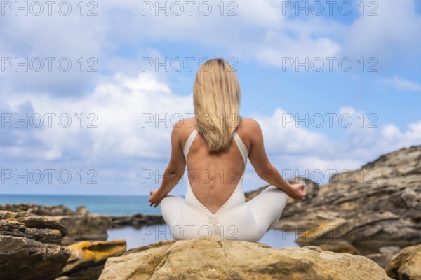 Woman in yoga attire seated in lotus on a coastal rock, meditating with calm focus amid blue ocean and clear sky, embodying balance, wellness, and serene outdoor mindfulness