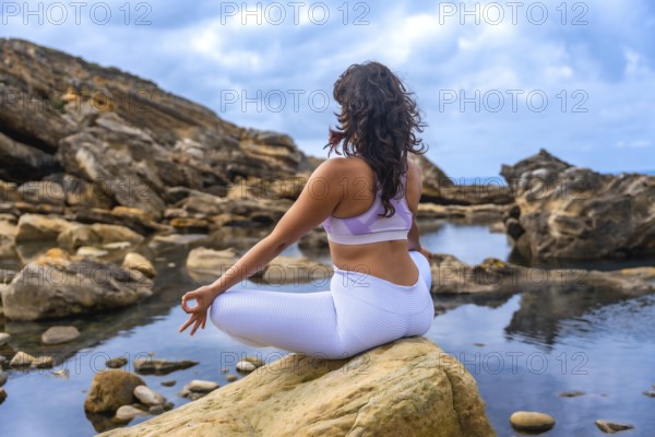 Woman performing a yoga meditation pose by a natural rock pool, sitting on a rock in a serene coastal landscape, finding calm and mindfulness in nature