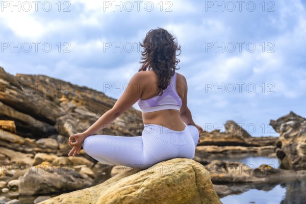 Woman sitting in a yoga lotus position on a rock, practicing meditation and mindfulness in a natural outdoor setting with rocky terrain and water pools under a cloudy sky