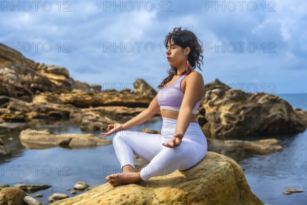 Woman sitting in a yoga pose, meditating outdoors on a rock by the sea, finding tranquility and balance amidst the natural coastal environment on a cloudy day