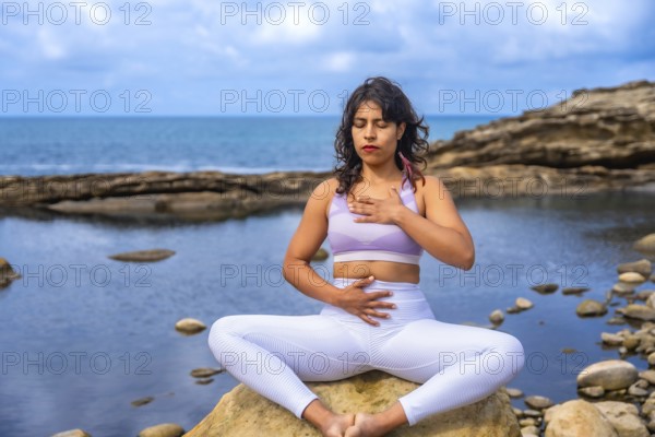 Woman practicing mindfulness and holistic wellness, sitting in a meditative yoga pose on a rock at the rocky coast, connecting with nature and focusing on mindful breathing