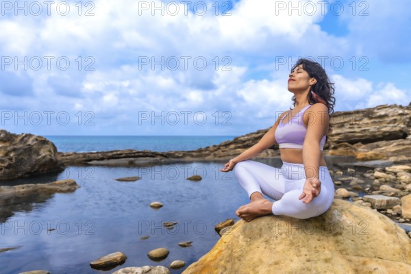 Woman in sportswear practicing yoga and meditation in lotus position on a large rock by the sea, finding inner peace and wellness in a serene natural environment