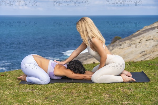 Two women are practicing yoga on a mat on a grassy cliff overlooking the ocean, with one woman gently guiding another in a relaxing child's pose, focusing on wellness and mindfulness