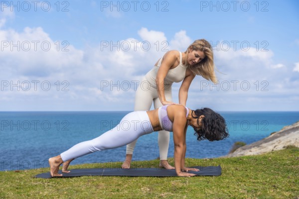 Female yoga instructor assisting a woman in plank pose on a mat, emphasizing proper alignment and technique during an outdoor wellness class by the ocean