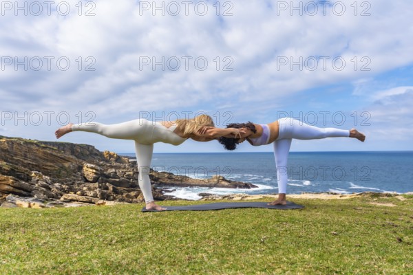 Two women practicing acro yoga outdoors on a grassy cliff overlooking the ocean, holding a deep warrior pose together, symbolizing balance, teamwork, and wellness