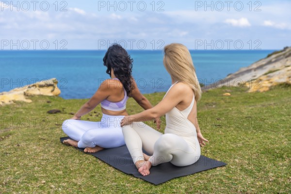 Two diverse women performing a seated spinal twist yoga pose on mats, enjoying a serene outdoor fitness and wellness session by the blue ocean and green cliff