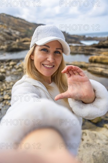 Blonde woman in white cap and jacket smiling at camera on a rocky beach, forming a heart with her handspeaceful, joyful portrait conveying self love and seaside wellness