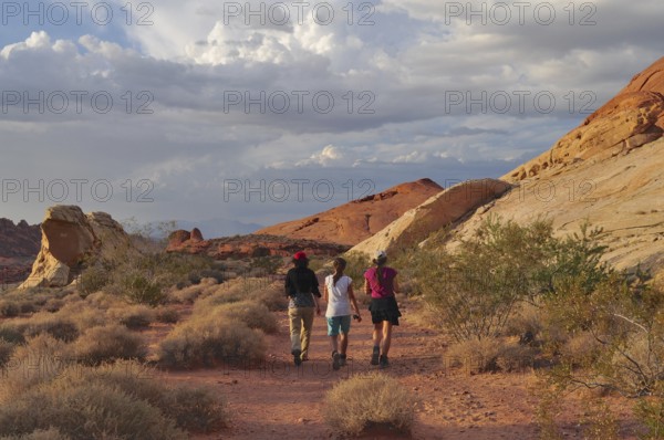 Three hikers on a trail through a desert landscape with clouds, Valley of Fire State Park, Nevada, USA