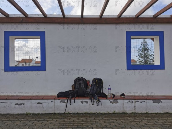 Two backpacks on a bench in front of a white wall with blue window cutouts, hiking on the Fisherman's Trail, Algarve, Portugal