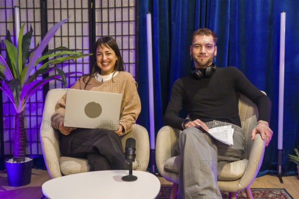 Two smiling content creators, a woman holding a laptop and a man holding notes, sitting in chairs during a podcast or social media recording session in a modern studio