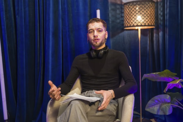 Young man wearing headphones around his neck, holding papers, and gesturing while speaking, actively creating content for social media or recording a podcast in a professional studio setup