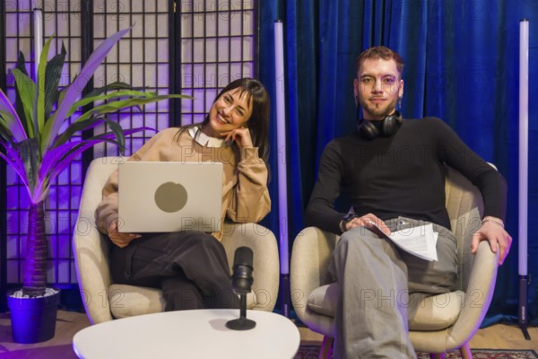 Two young content creators, a smiling woman with a laptop and a man with headphones, sitting on chairs and preparing for a live podcast recording in a modern studio setup