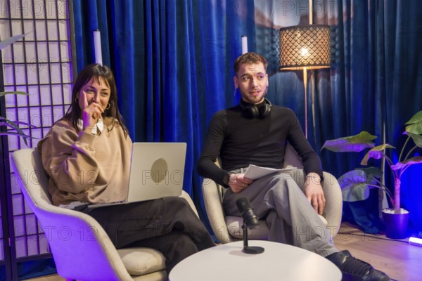 Two young content creators engaging in a podcast recording session, using a laptop, microphone, and headphones within a vibrant studio setup for social media and streaming
