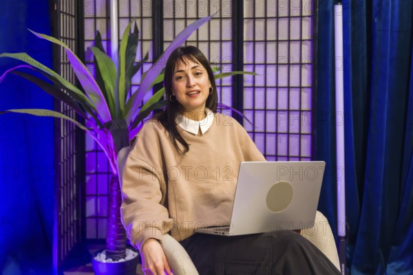 Woman sitting in a professional content creation studio, holding a laptop in her lap while looking towards the camera, ready for recording podcasts or social media videos