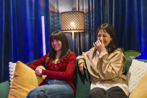 Two young women sitting on a sofa, one smiling and the other laughing, collaborating creatively in a modern content creation studio for podcasts and online platforms