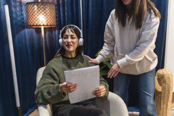 Young woman in headphones holding papers and smiling while preparing to record a podcast or social media content in a modern studio, assisted by a colleague