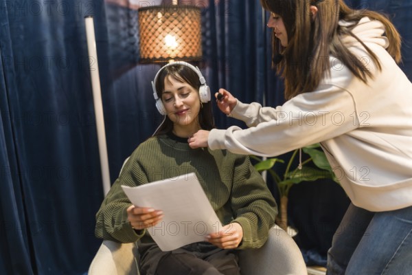 Content creator preparing for podcast recording in a professional studio, wearing headphones and holding a script while a sound technician adjusts her lavalier microphone