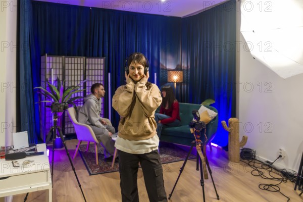 Young woman smiling and wearing headphones while standing in a trendy podcast and content creation studio with friends and professional equipment, ready for digital media production