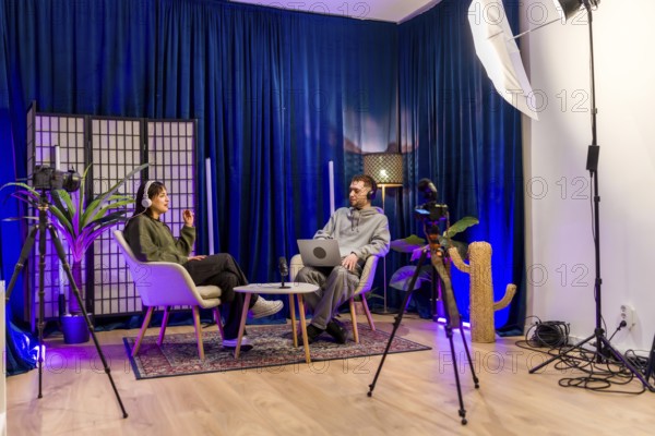 Two young adults in a professional podcast studio record an interview with headphones, laptop, cameras and lighting, creating social media and video content together
