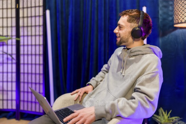 Young man creating digital content, recording a podcast or live stream, smiling and using a laptop while wearing headphones in a modern studio setting with blue and purple lighting