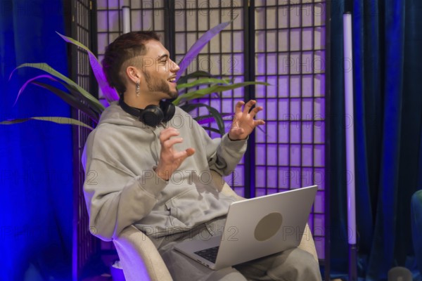 Young man wearing headphones and using a laptop, actively gesturing while recording a podcast or live streaming social media content in a modern studio