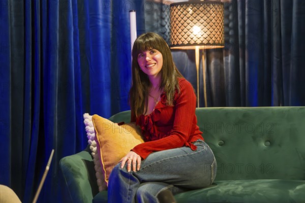 Young smiling woman sitting on a green couch in a contemporary recording studio, engaged in content creation for social media or a podcast, with a microphone and unique lighting