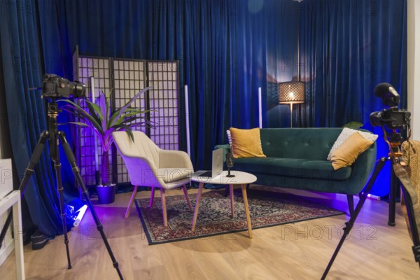Modern content creation studio equipped for podcast recording or social media video production, featuring cameras on tripods, a microphone, laptop, sofa, armchair, and blue velvet curtains
