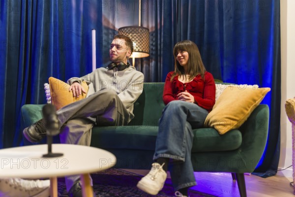 Young man and woman sitting on a green sofa, actively listening and engaging while recording a podcast or digital content in a modern studio setup with a microphone on a coffee table