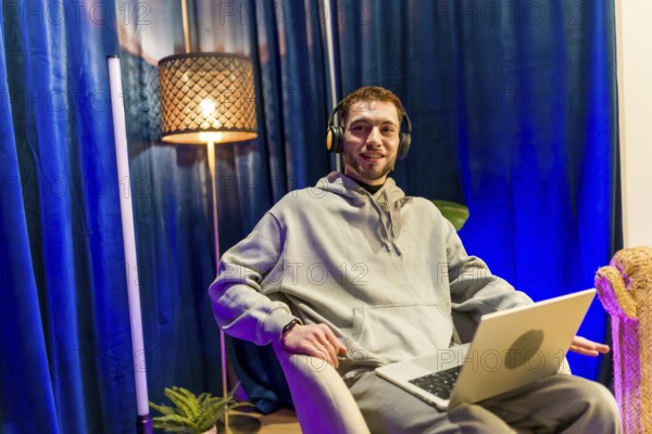Young man wearing headphones and hoodie, sitting comfortably with a laptop in a home studio, smiling while streaming or recording a podcast for social media content
