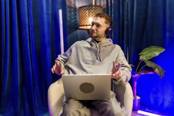 Young man wearing headphones and using laptop, recording a podcast or live streaming content from his creative home studio with colorful ambient lighting