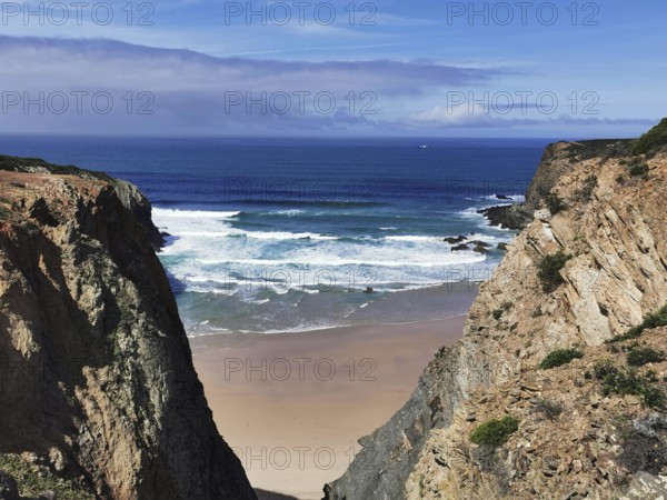 Narrow view of the beach between two rocky cliffs with blue sea and slightly cloudy sky, hiking on the Fisherman's Trail, Algarve, Portugal